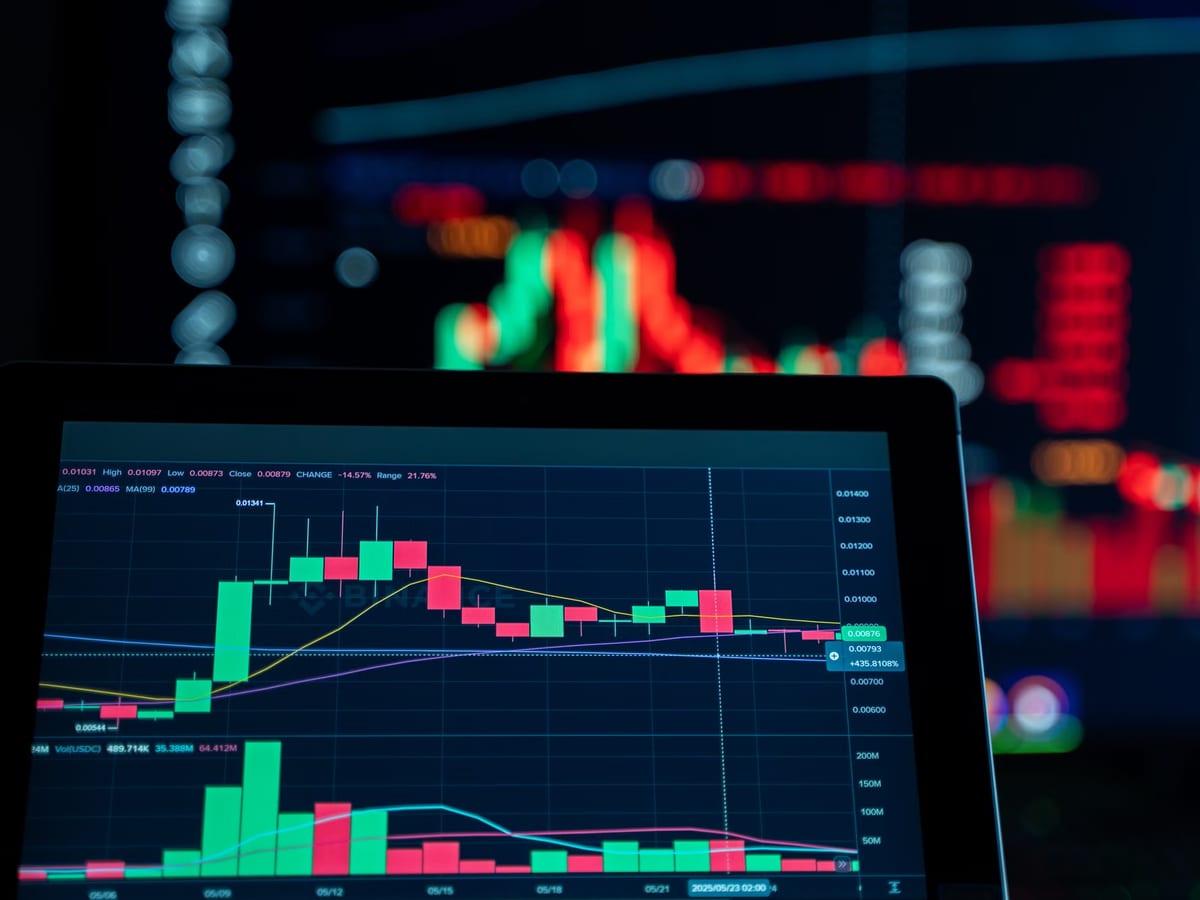 A laptop and monitor showing candlestick price charts in a darkened room