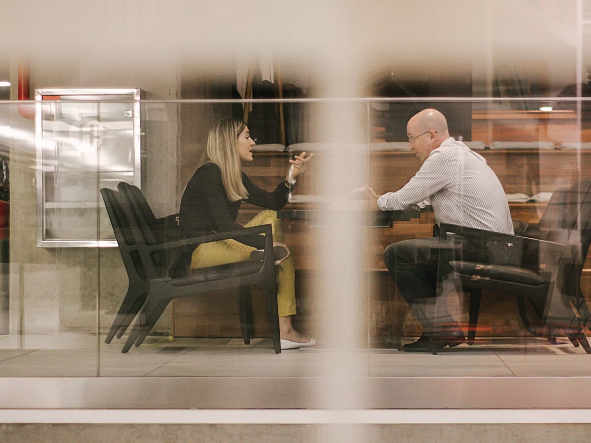 Two colleagues in conversation in a glass-walled meeting room, seen through a reflective partition
